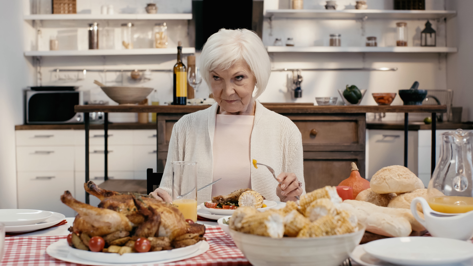 An elderly woman with short white hair sits at a dining table set with various foods, including a roast chicken, corn on the cob, bread, and salad. She is holding a fork and looking off to the side, with a kitchen in the background.