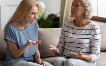 A young woman and an older woman sit on a beige couch engaged in a serious conversation. The younger woman, with blonde hair, gestures with her hand while looking intently. The older woman, with gray hair, responds thoughtfully. Both appear focused and concerned.