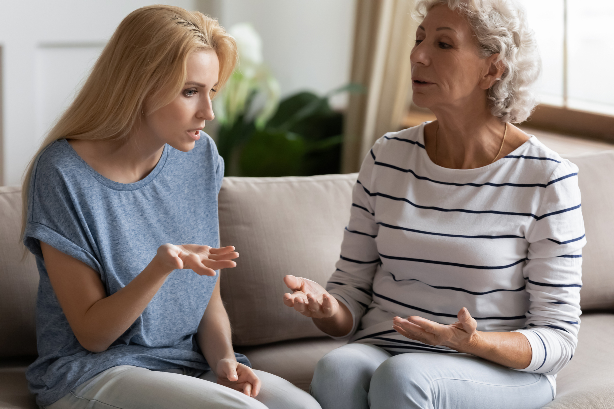 A young woman and an older woman sit on a beige couch engaged in a serious conversation. The younger woman, with blonde hair, gestures with her hand while looking intently. The older woman, with gray hair, responds thoughtfully. Both appear focused and concerned.