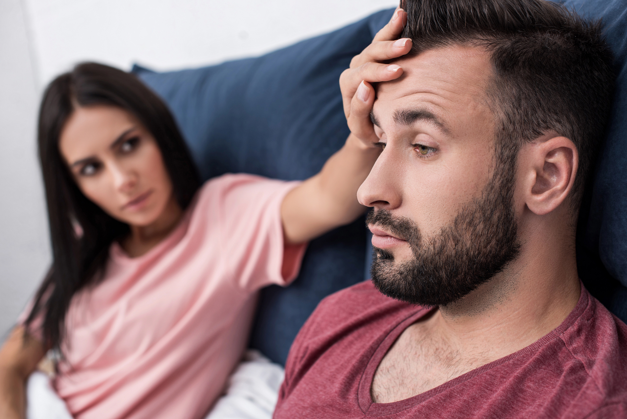 A woman in a pink shirt is looking concerned as she rests her hand on the forehead of a man in a maroon shirt, who appears deep in thought. They are sitting on a dark blue couch, with the woman leaning towards the man.