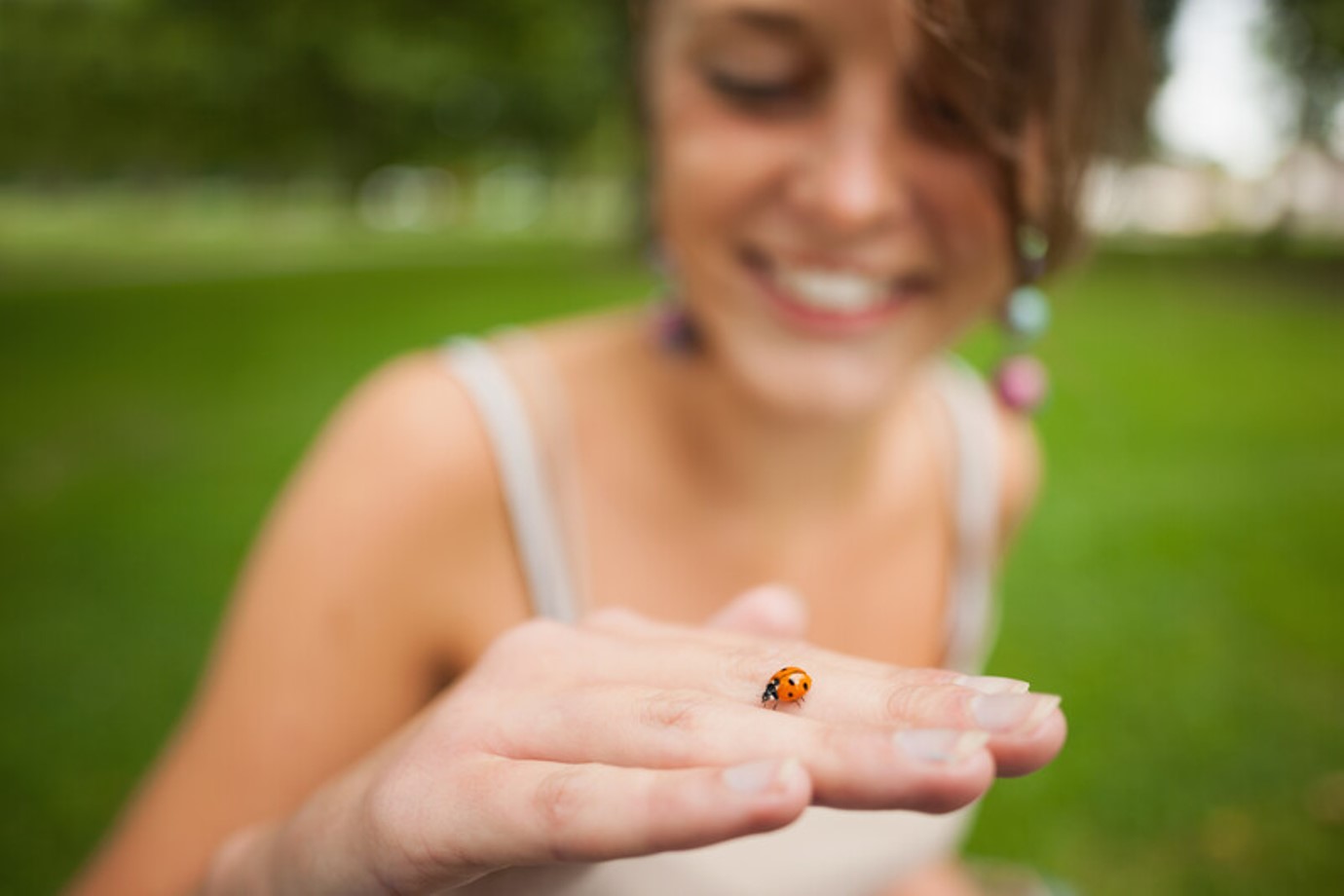 A smiling person holds their hand up close to the camera, with a small ladybug crawling on their finger. The background is a blurry green outdoor setting.