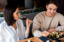 A woman in a white shirt is drinking red wine while sitting at a restaurant table with a man in a beige sweatshirt who is using chopsticks to eat sushi. Several pieces of sushi and a small dish are on the table in front of them.