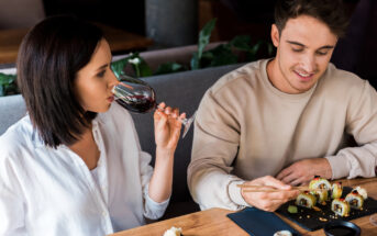 A woman in a white shirt is drinking red wine while sitting at a restaurant table with a man in a beige sweatshirt who is using chopsticks to eat sushi. Several pieces of sushi and a small dish are on the table in front of them.