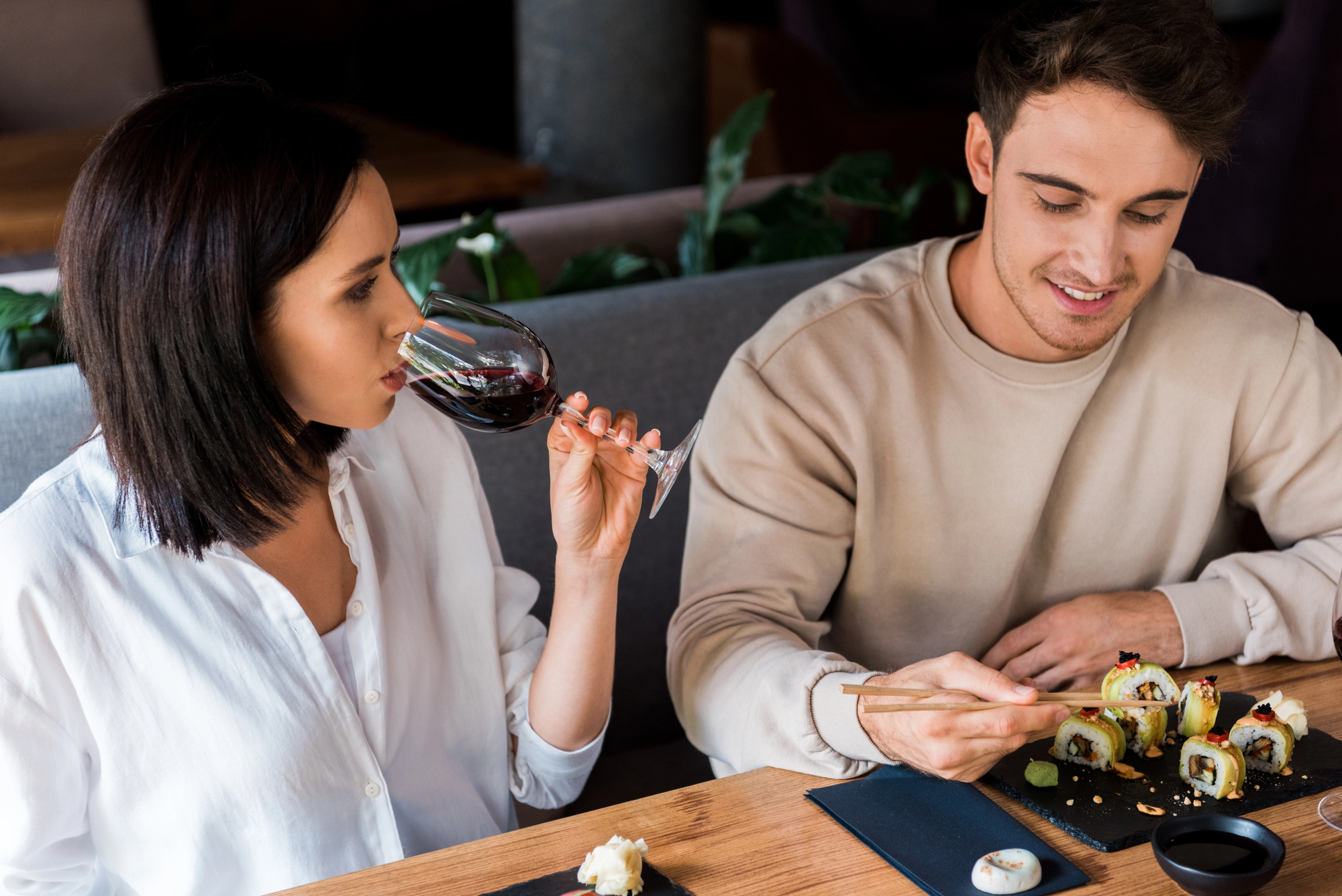 A woman in a white shirt is drinking red wine while sitting at a restaurant table with a man in a beige sweatshirt who is using chopsticks to eat sushi. Several pieces of sushi and a small dish are on the table in front of them.