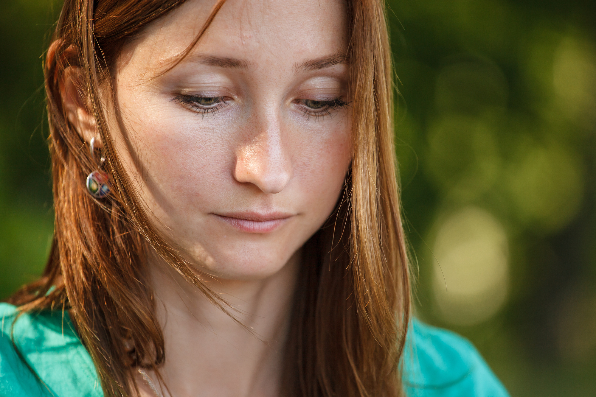 A woman with long auburn hair looks down pensively. She is wearing green clothing and earrings, with a blurred natural background of greenery. The sunlight softly illuminates her face.