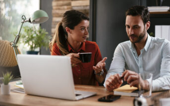 A woman in a red shirt holds a coffee cup and gestures while talking to a man in a light blue shirt. They are seated at a wooden desk with a laptop, a smartphone, and a glass of water. The workspace is decorated with plants and has a modern aesthetic.