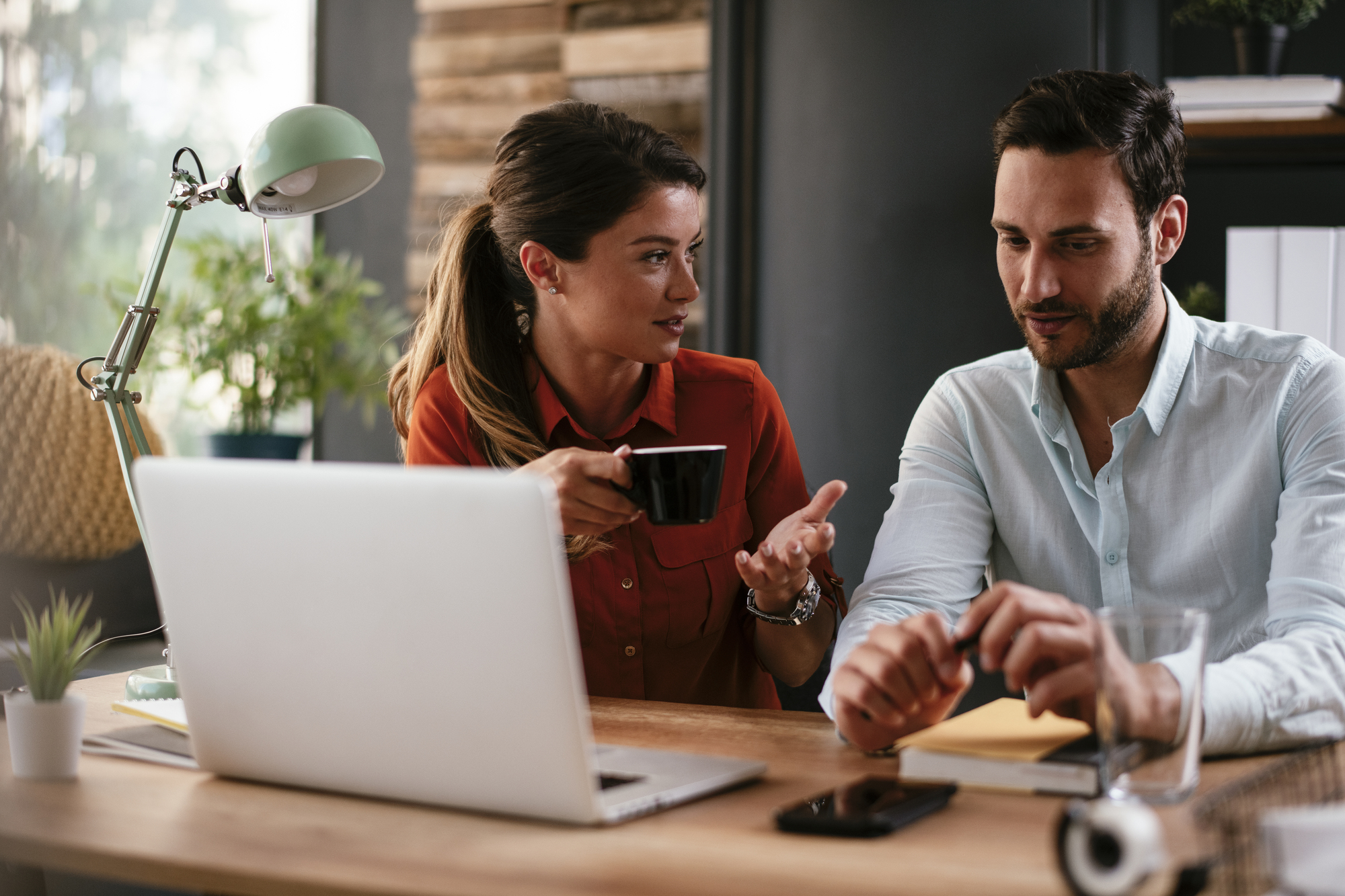 A woman in a red shirt holds a coffee cup and gestures while talking to a man in a light blue shirt. They are seated at a wooden desk with a laptop, a smartphone, and a glass of water. The workspace is decorated with plants and has a modern aesthetic.