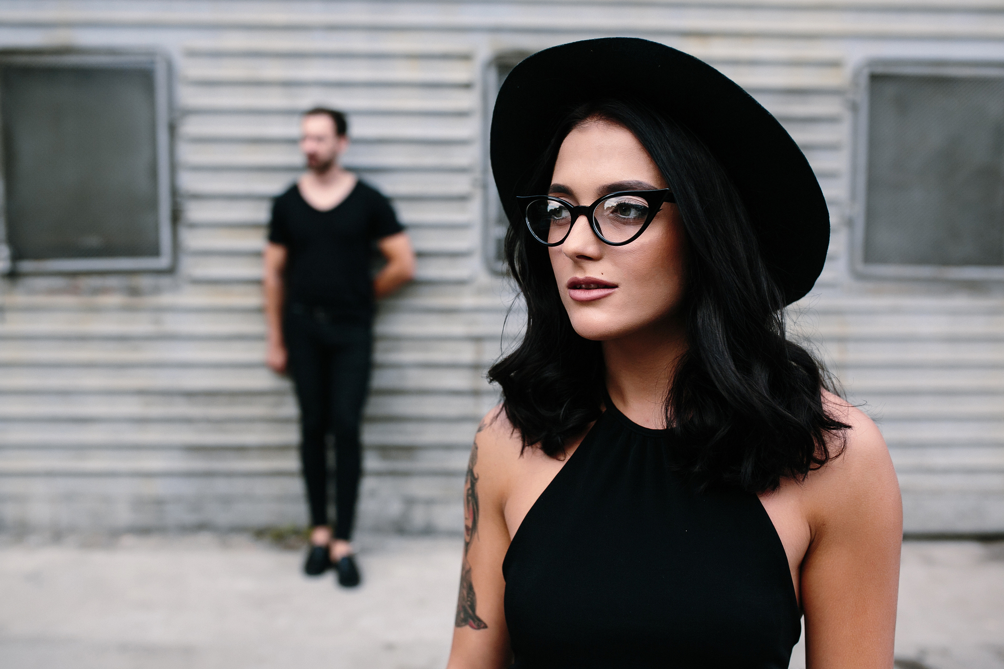 A woman with shoulder-length dark hair, wearing a black hat, black-framed glasses, and a sleeveless black top, stands in the foreground looking to the side. In the blurred background, a man in black clothing stands against a corrugated metal wall.