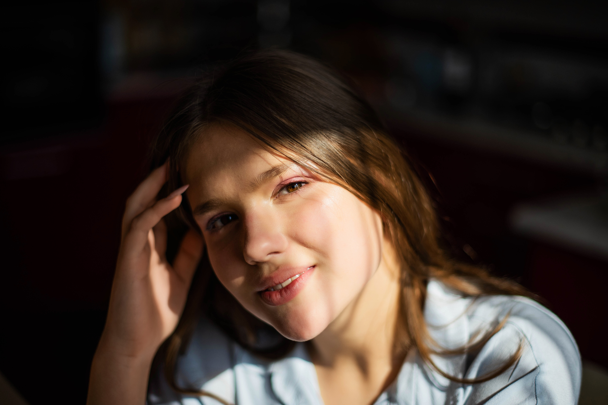 A young person with long brown hair smiles gently while resting their hand on the side of their head. They are bathed in soft sunlight, creating a warm and inviting atmosphere. The background is blurred, focusing attention on their face.