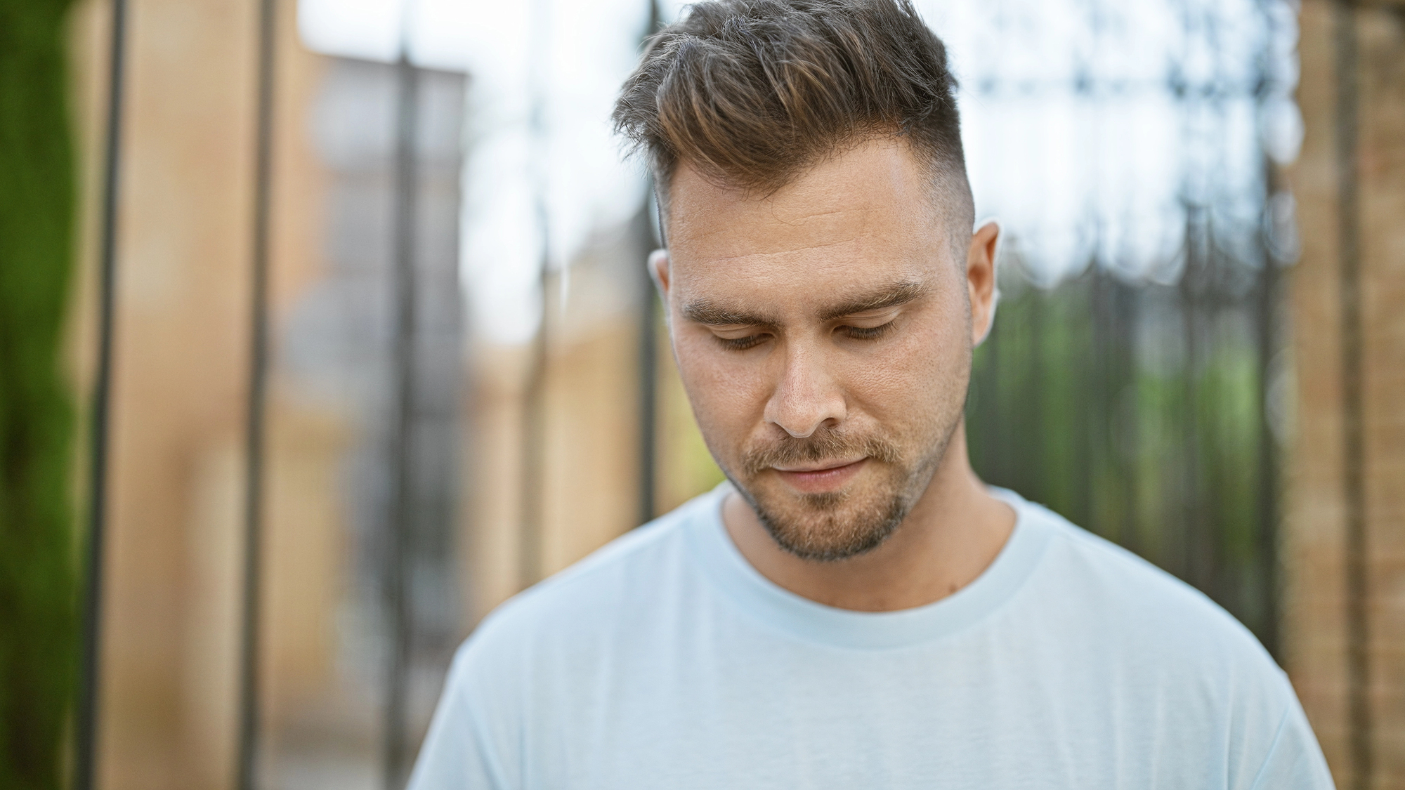 A man with short, styled hair and a trimmed beard looks down with a neutral expression. He is wearing a light blue t-shirt and standing outside, with a blurred background featuring a building and a fence.