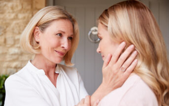 A woman with short blonde hair gently holds the face of a younger woman with long blonde hair, both looking at each other lovingly. They are in front of a light grey door with a circular knocker. The scene captures a tender moment of connection.