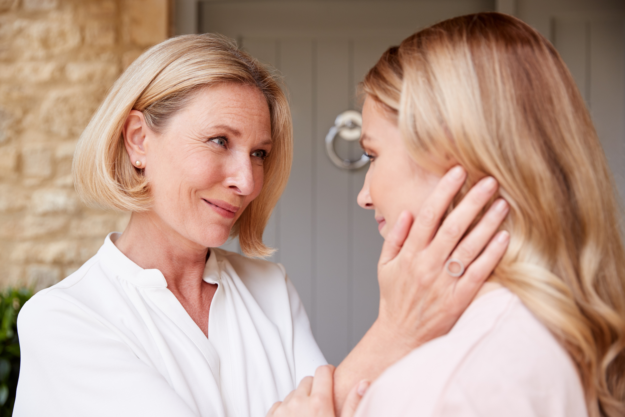 A woman with short blonde hair gently holds the face of a younger woman with long blonde hair, both looking at each other lovingly. They are in front of a light grey door with a circular knocker. The scene captures a tender moment of connection.