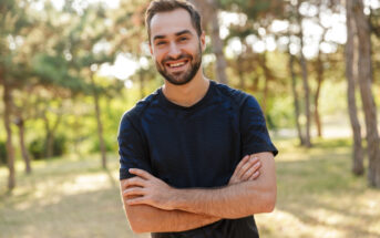 A man with a beard and short hair is smiling and standing outdoors with his arms crossed. He is wearing a dark patterned shirt. The background features trees and greenery, suggesting a park or forest area, with sunlight filtering through the foliage.