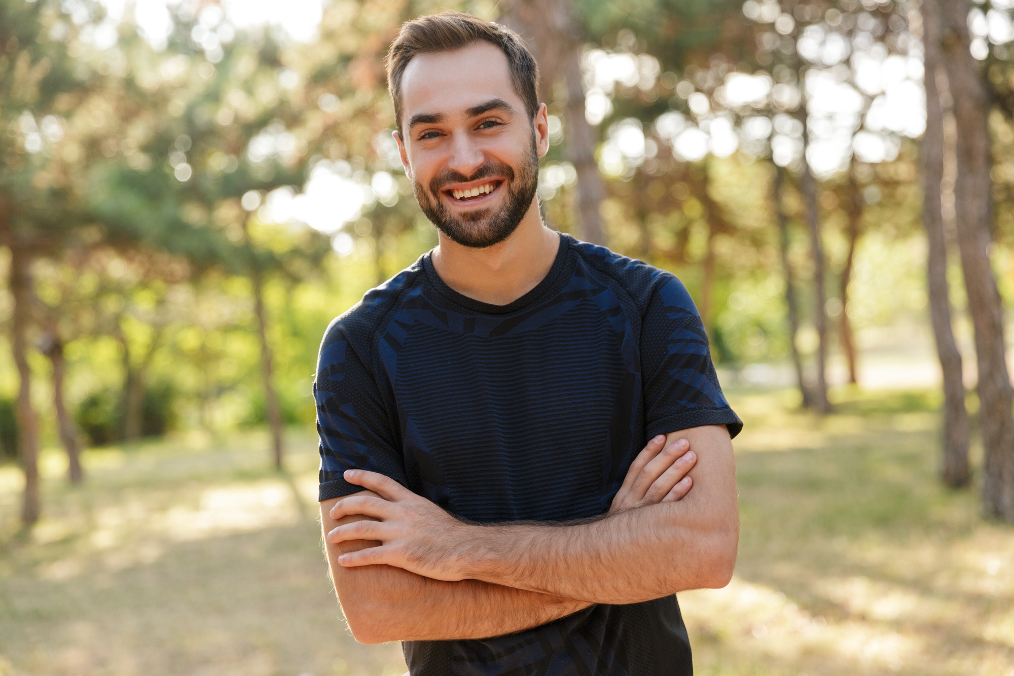 A man with a beard and short hair is smiling and standing outdoors with his arms crossed. He is wearing a dark patterned shirt. The background features trees and greenery, suggesting a park or forest area, with sunlight filtering through the foliage.