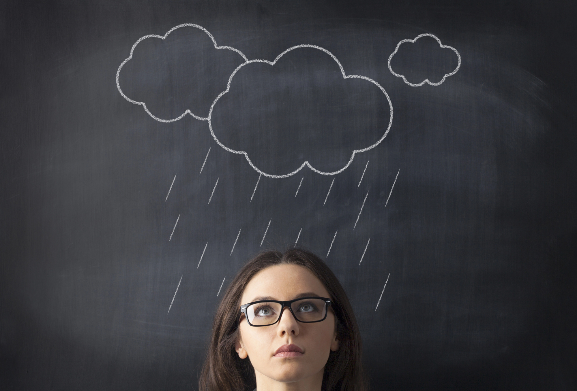 A woman wearing glasses looks up with a neutral expression, standing in front of a blackboard with chalk drawings of clouds and rain above her head, suggesting a metaphor for thinking or feeling under the weather.