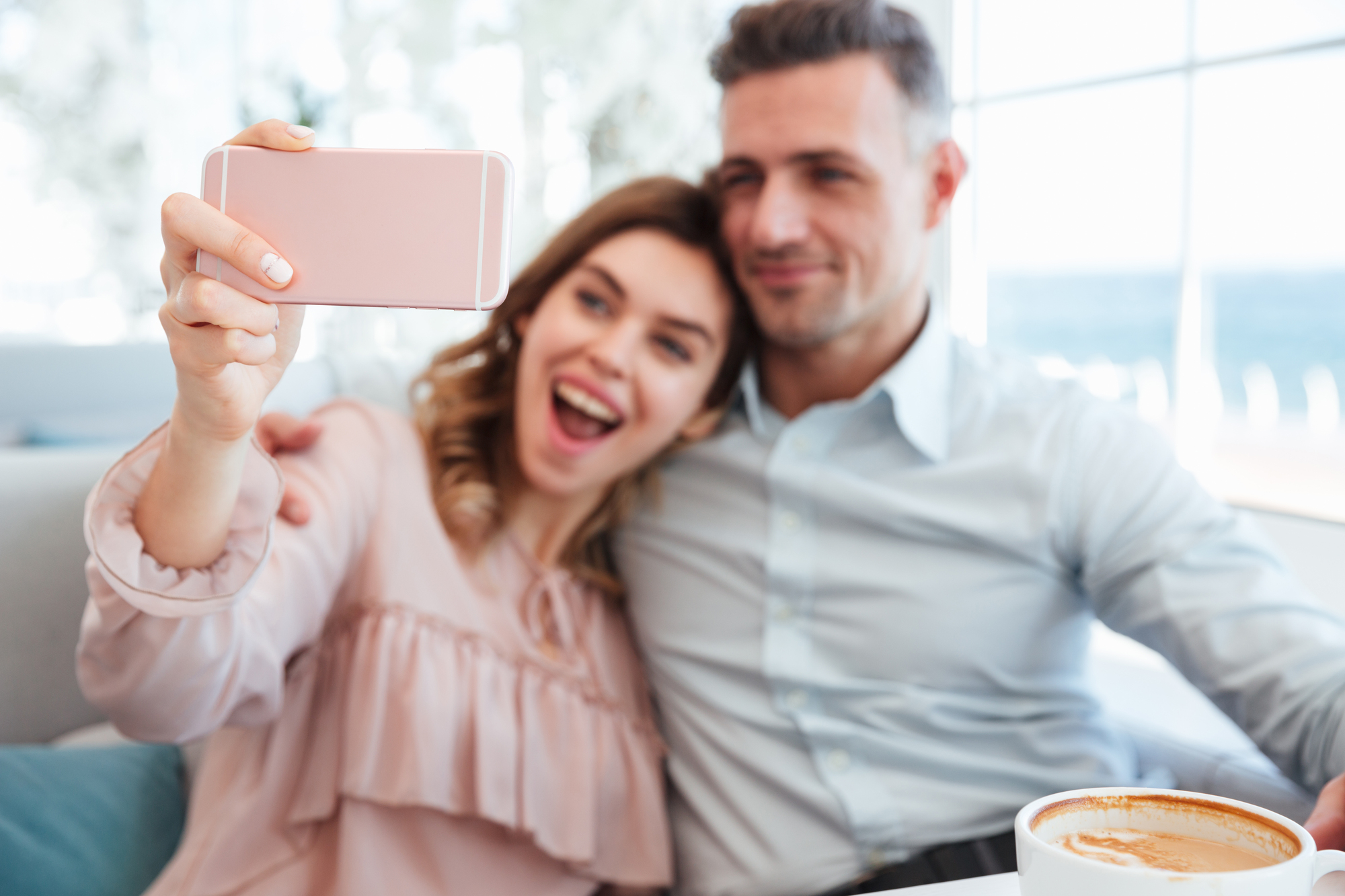 A smiling woman and a man sitting closely together in a bright café, taking a selfie with a pink smartphone. The woman has shoulder-length hair and is wearing a pale pink blouse. The man has short hair and is wearing a light blue shirt. A coffee cup is visible in the foreground.