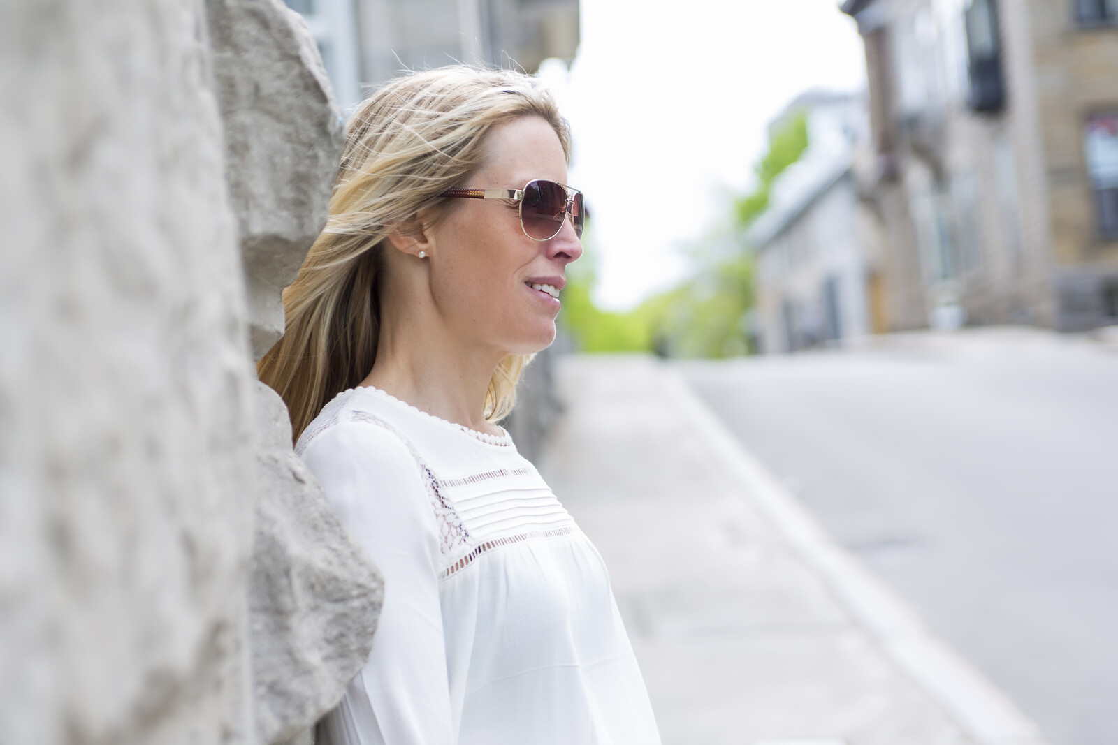 A woman with long blonde hair and sunglasses leans against a stone wall, smiling and looking to the side. She is wearing a white blouse, and the background features a street with blurred buildings and greenery.