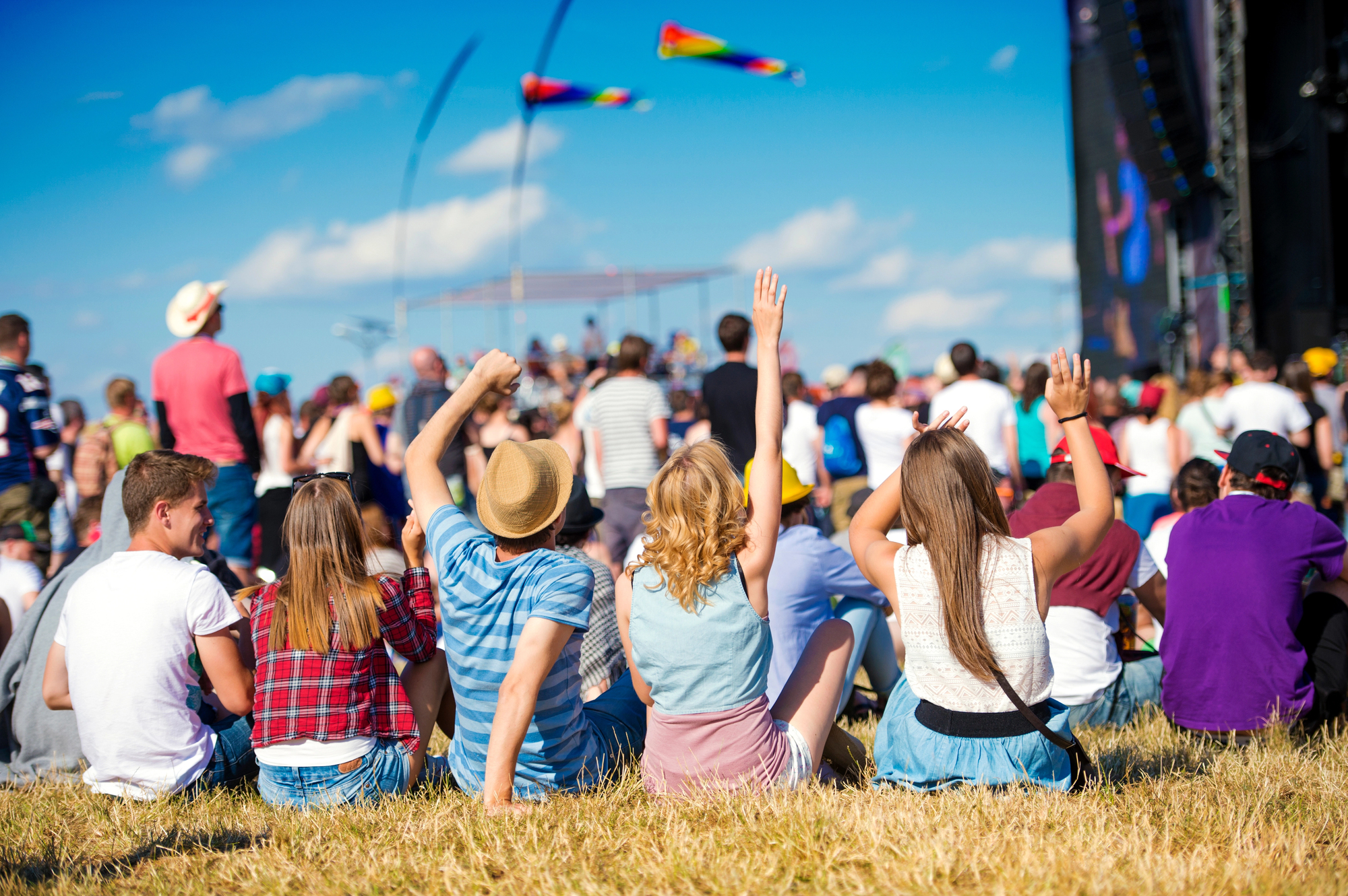 A group of young people sits on grass, enjoying an outdoor music festival or concert on a sunny day. Some raise their arms, while others chat. The background shows a large stage with performers and colorful flags flying in the sky.