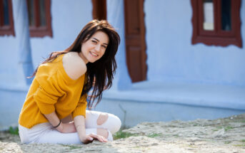 A woman with long dark hair is sitting outside on a rocky surface, smiling at the camera. She is wearing a yellow off-shoulder sweater and white ripped jeans. Behind her, there is a blue building with red window frames and a brown door.