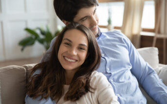 A smiling woman with long brown hair leans back on a couch, resting her head on the chest of a man with dark hair who is sitting behind her. The man has his arm around her, and both appear relaxed and happy in a cozy, well-lit room.