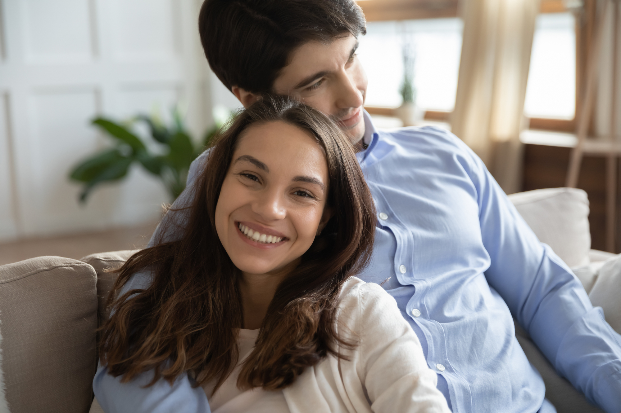 A smiling woman with long brown hair leans back on a couch, resting her head on the chest of a man with dark hair who is sitting behind her. The man has his arm around her, and both appear relaxed and happy in a cozy, well-lit room.