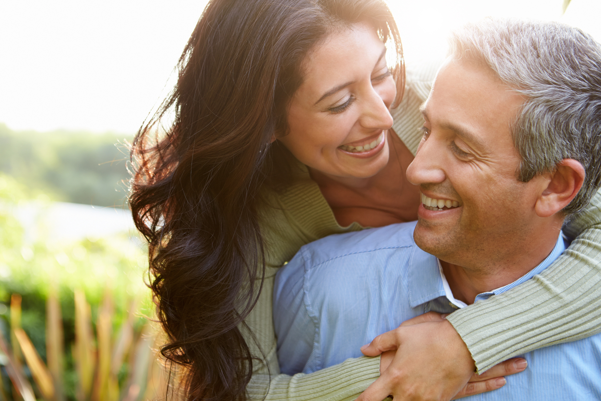 A woman with long, dark hair is hugging a man with short, graying hair from behind. They are both smiling and looking at each other, standing outdoors with greenery and sunlight in the background.