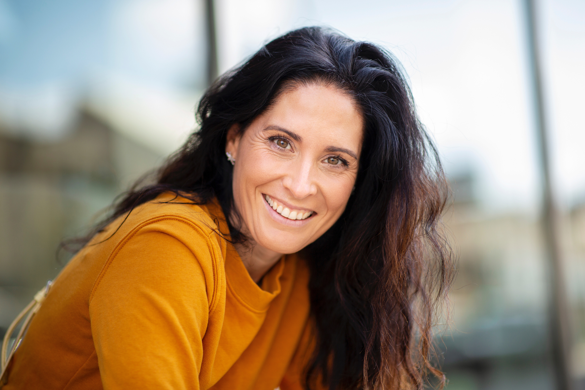 A woman with long, dark hair is smiling at the camera. She is wearing a mustard yellow top and is outdoors with a blurred background, likely an urban setting. The image exudes a friendly and approachable vibe.