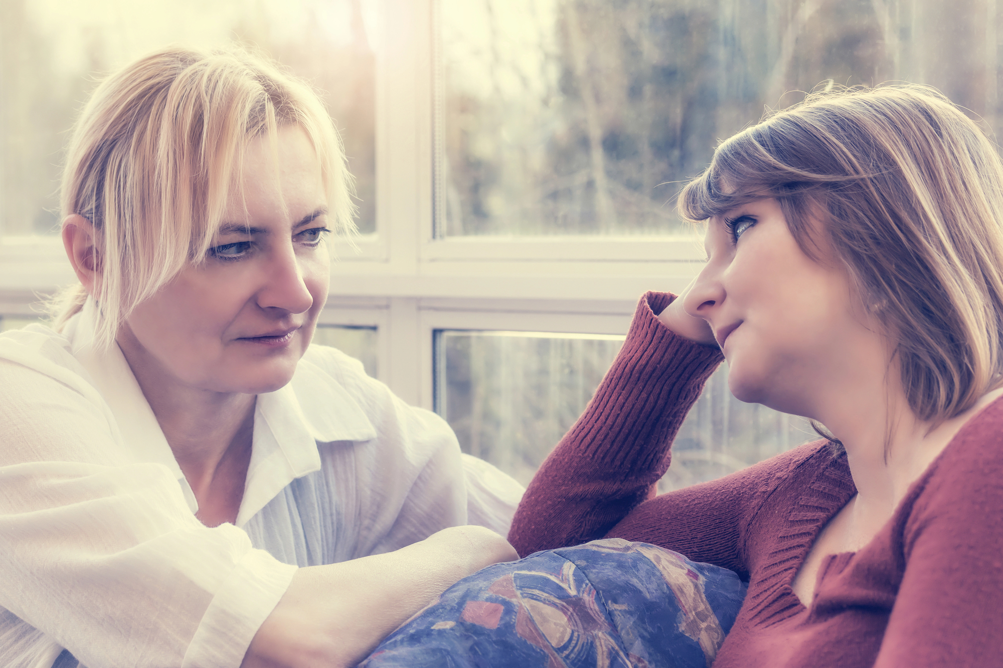 Two women sitting close to each other, engaged in a deep conversation. The woman on the left, wearing a white blouse, appears thoughtful. The woman on the right, in a rust-colored sweater, leans on a cushioned surface, gazing intently at her companion.