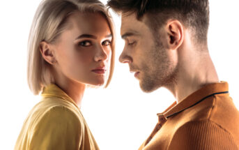 A close-up of a young woman with short blonde hair and a serious expression, wearing a yellow top, looking toward the camera. Next to her, a young man with short brown hair in an orange polo shirt looks down and away from the camera against a white background.