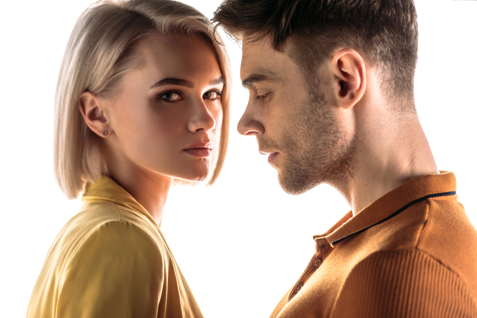 A close-up of a young woman with short blonde hair and a serious expression, wearing a yellow top, looking toward the camera. Next to her, a young man with short brown hair in an orange polo shirt looks down and away from the camera against a white background.