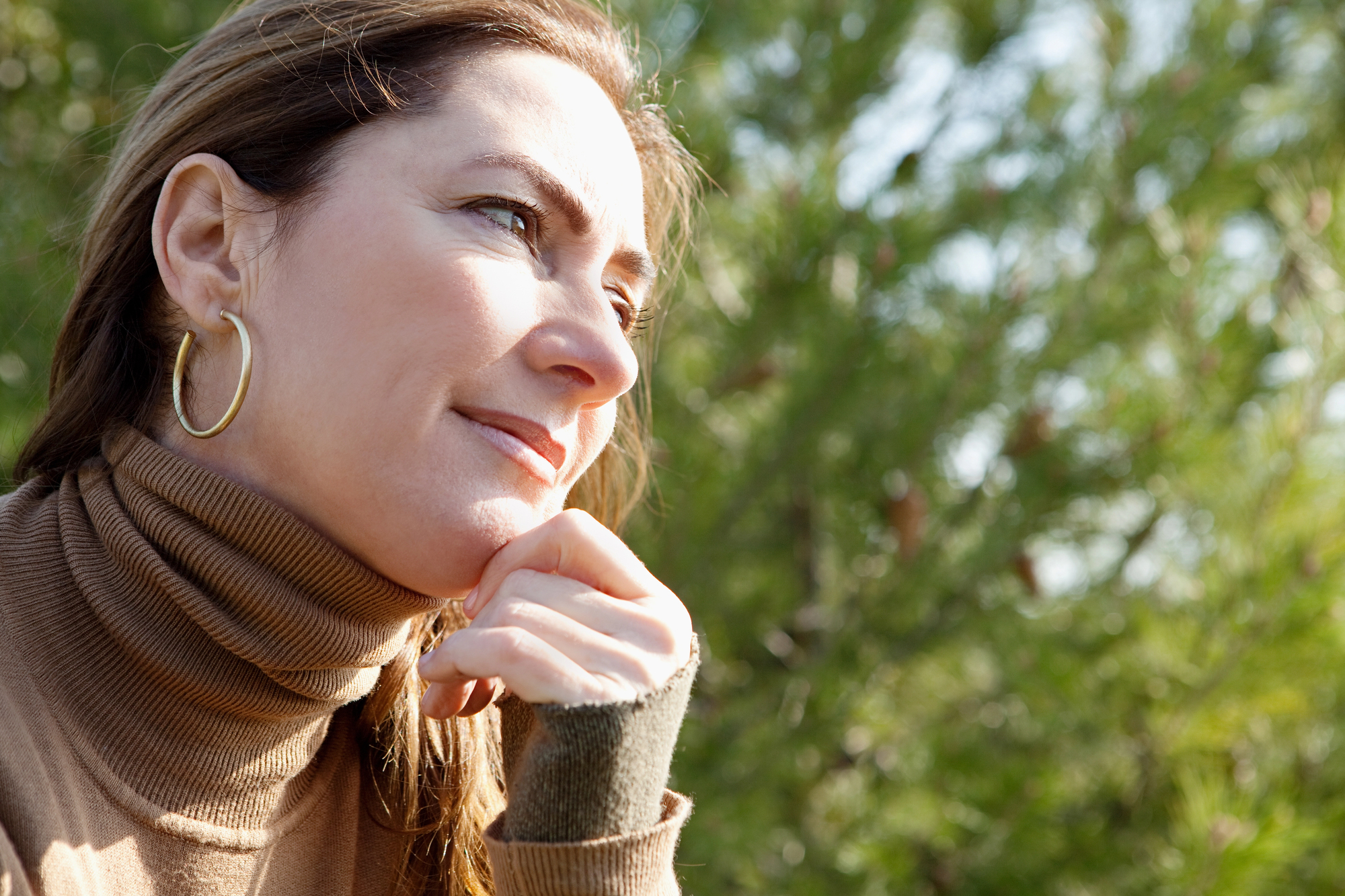 A woman with light brown hair wearing a brown turtleneck sweater and gold hoop earrings is seen in a side profile shot. She is resting her chin on her hand and looking thoughtfully into the distance with a backdrop of lush green foliage.