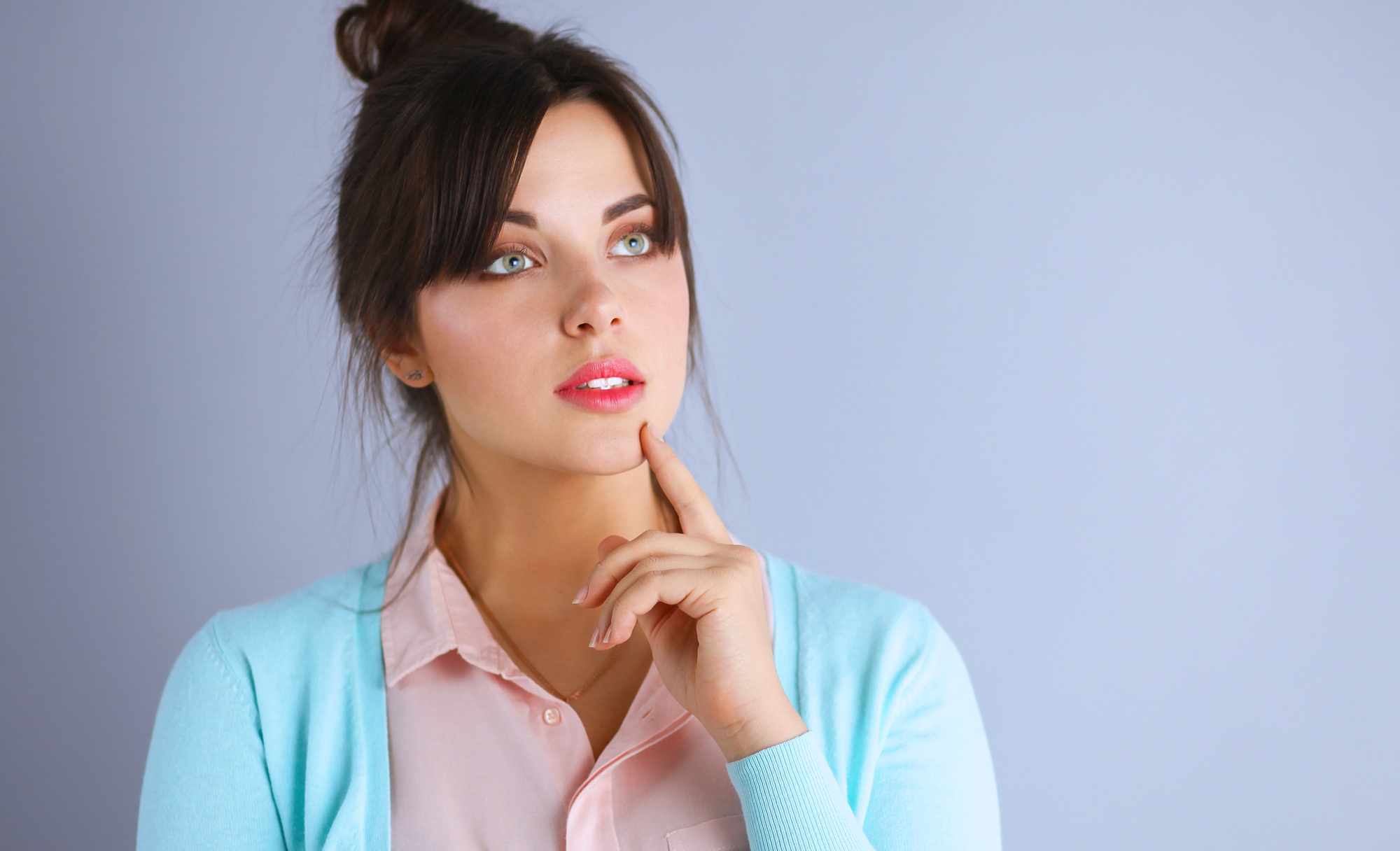 A woman with dark hair tied in a bun is standing against a light gray background. She is looking upwards pensively with her index finger resting on her chin. She is wearing a light blue sweater over a pink blouse.