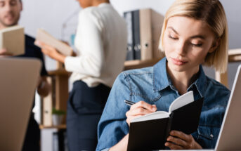 A woman with short blonde hair is focused on writing in a small notebook while sitting at a desk with a laptop in front of her. In the background, two people are standing and talking, with a shelf of binders and books behind them. The setting appears to be an office.