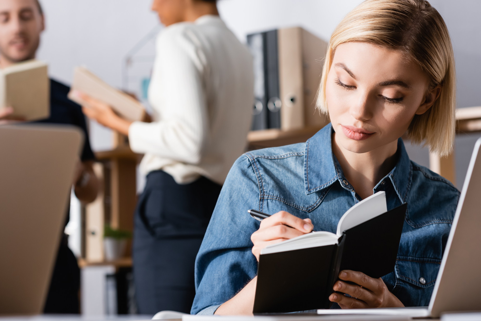 A woman with short blonde hair is focused on writing in a small notebook while sitting at a desk with a laptop in front of her. In the background, two people are standing and talking, with a shelf of binders and books behind them. The setting appears to be an office.