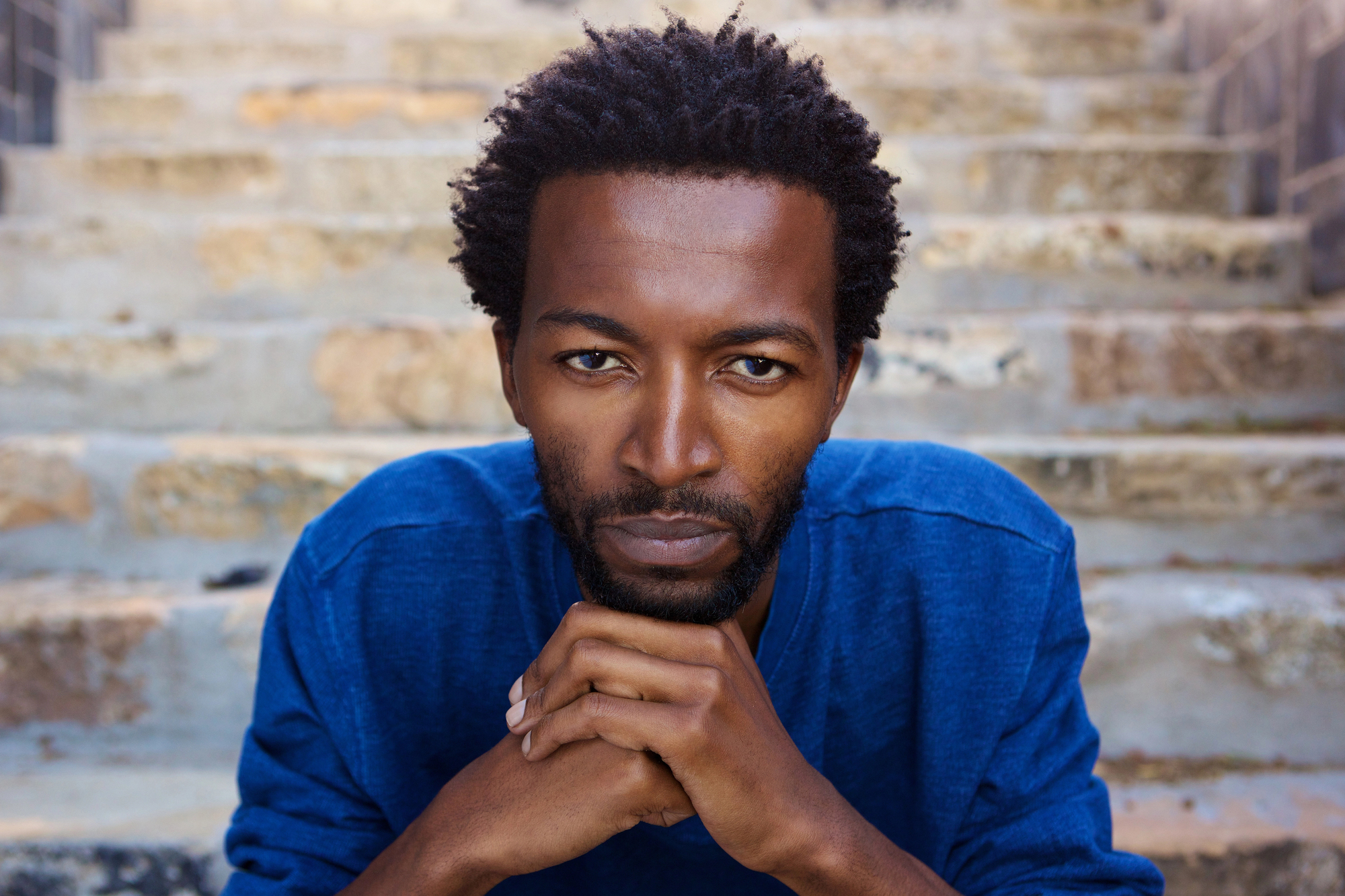 A man with short curly hair and a beard is resting his chin on his hands while sitting on stone steps. He is wearing a blue shirt and looking directly at the camera with a serious expression. The background consists of stone steps slightly out of focus.