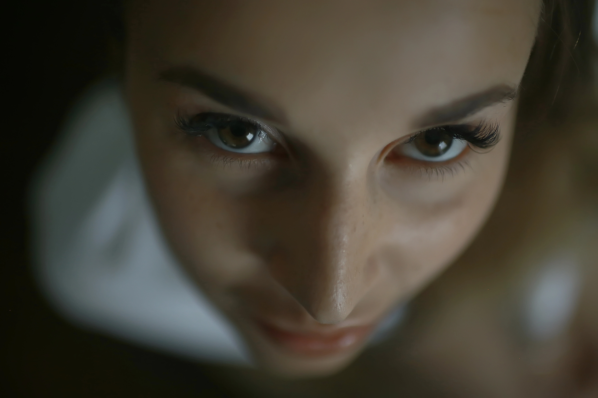 Close-up of a person's face with a focus on their eyes, which are looking directly at the camera. The person's gaze appears intense, with long eyelashes and a few freckles visible on their nose and cheeks. The background is dark and blurred.