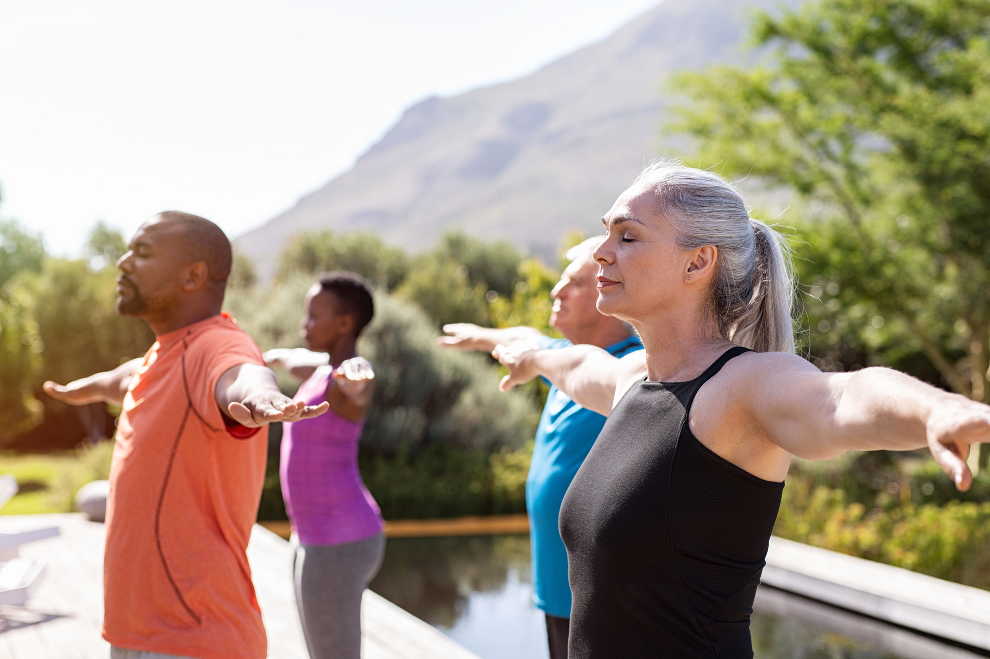 A diverse group of four adults, dressed in athletic wear, practice yoga outdoors. They are standing with eyes closed and arms extended, appearing serene. The background features lush greenery, a body of water, and mountain scenery under a clear sky.