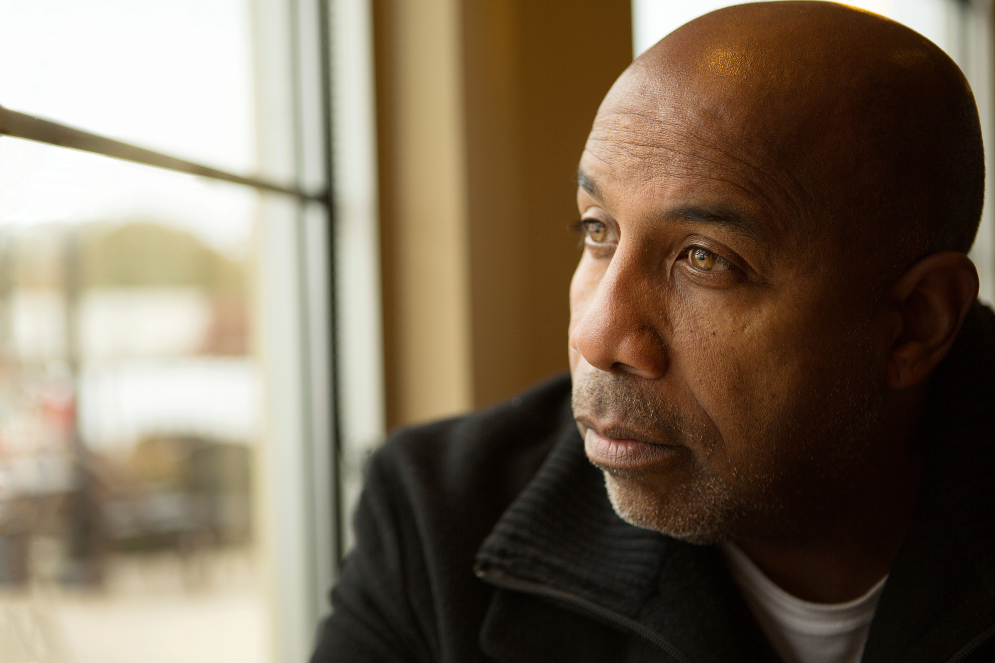 A man with a closely shaved head and light stubble gazes pensively out of a window. He is wearing a dark jacket and sitting inside a room with soft lighting. The blurred outdoor scene shows hints of a landscape beyond the window.