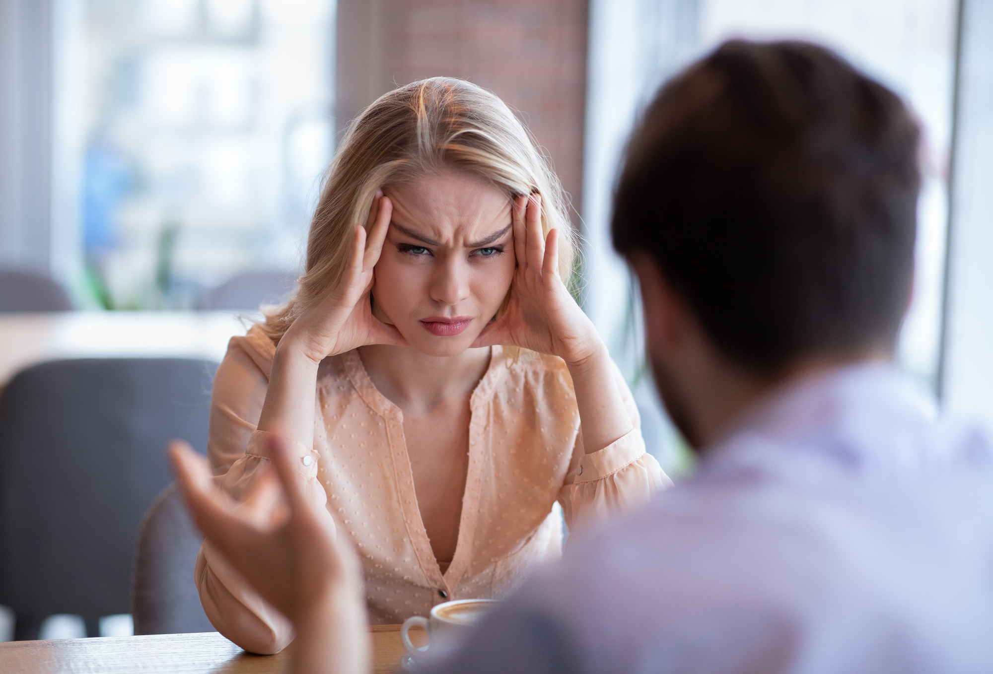 A woman with long blonde hair sits at a table in a coffee shop, looking stressed and resting her temples on her hands. She appears to be in a serious conversation with a man, who is facing away from the camera. Blurred background of the cafe interior.