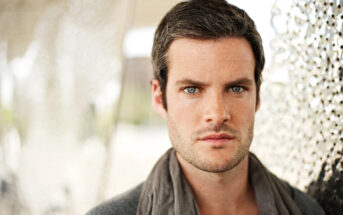 A man with short dark hair and light eyes looks directly at the camera with a serious expression. He is wearing a dark shirt and a light scarf. The background is slightly blurred with a mix of outdoor elements and patterned surfaces.