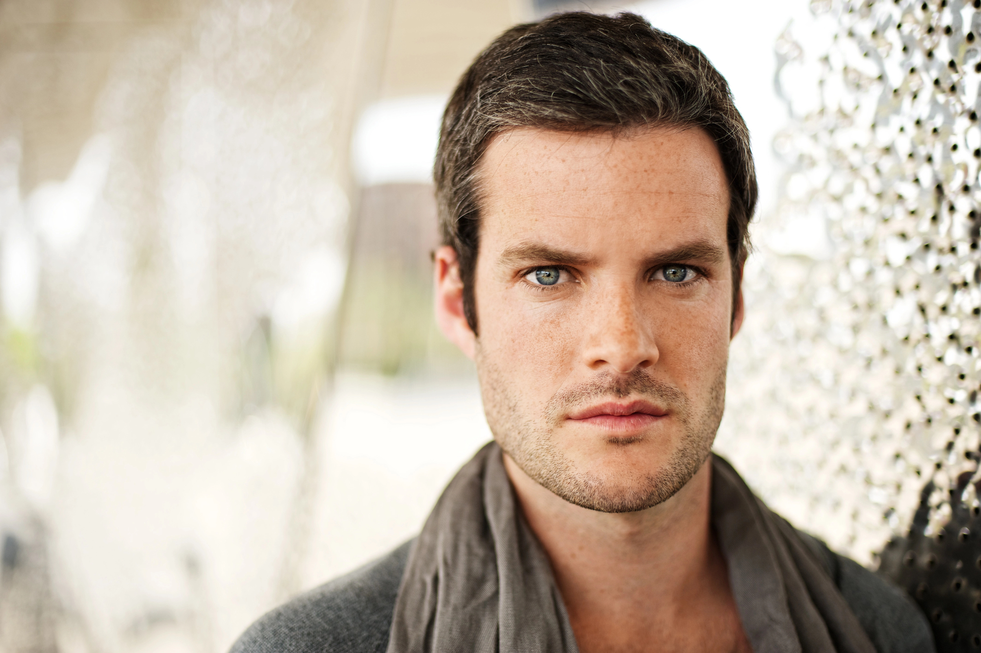 A man with short dark hair and light eyes looks directly at the camera with a serious expression. He is wearing a dark shirt and a light scarf. The background is slightly blurred with a mix of outdoor elements and patterned surfaces.