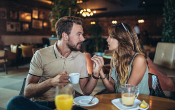 A man and a woman sit at a table in a cozy café, engaged in an animated conversation. The man holds a white coffee cup while the woman gestures with her hands. On the table are glasses of orange juice and an iced latte, along with a phone.