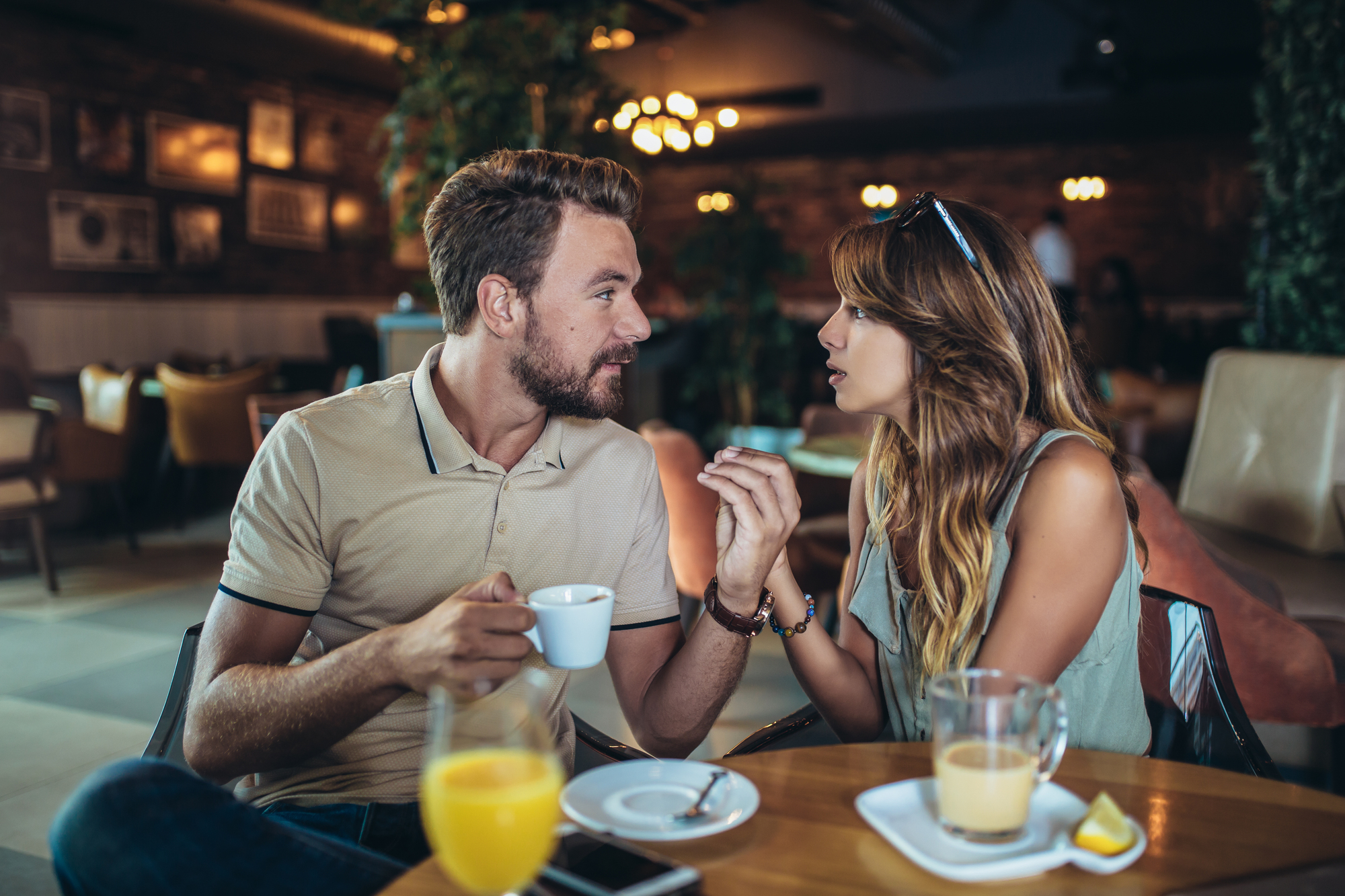 A man and a woman sit at a table in a cozy café, engaged in an animated conversation. The man holds a white coffee cup while the woman gestures with her hands. On the table are glasses of orange juice and an iced latte, along with a phone.