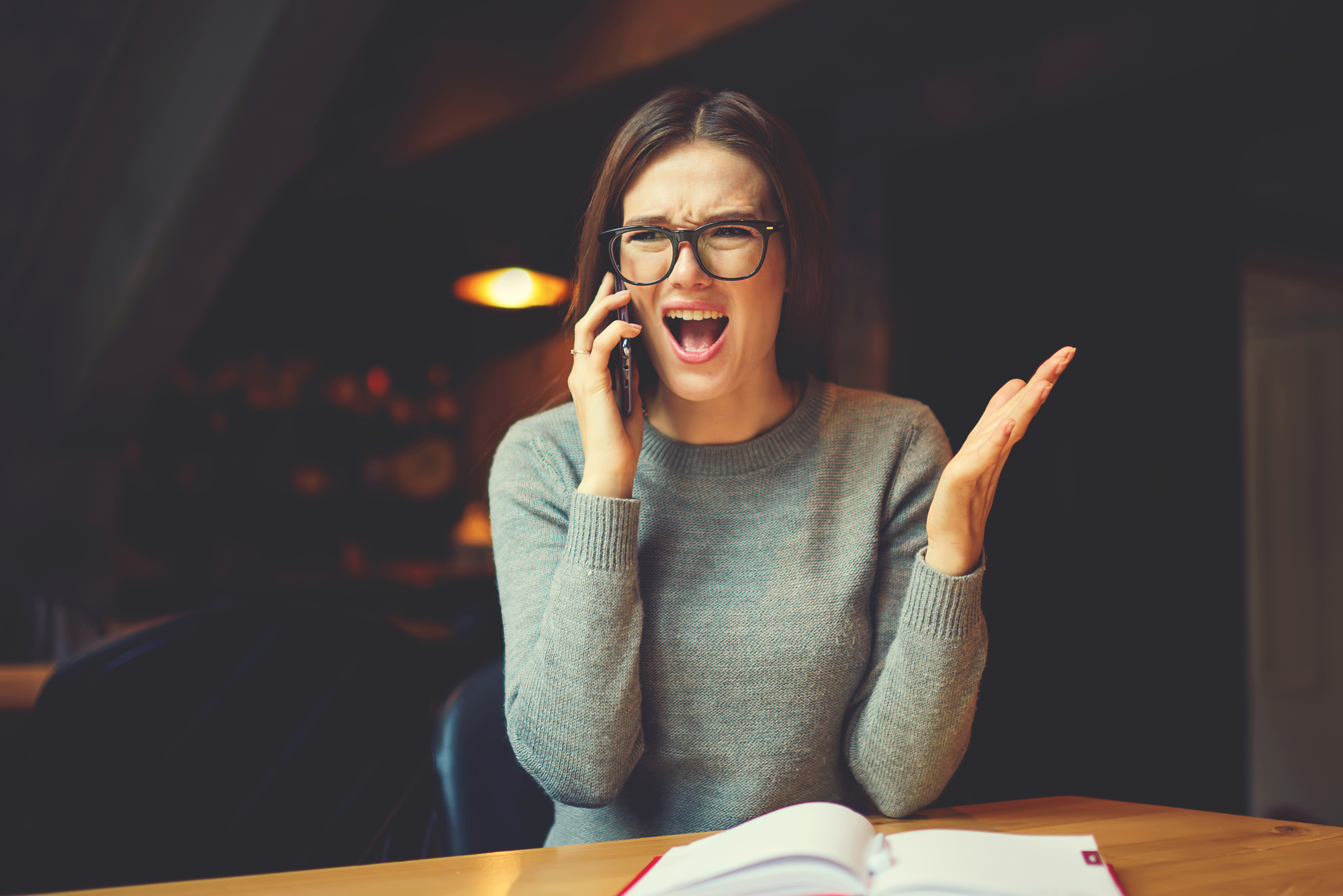 A woman with glasses is sitting at a wooden table, talking on a mobile phone with an open book in front of her. She appears to be frustrated or upset, raising her left hand with a distressed expression on her face. The background is dimly lit.