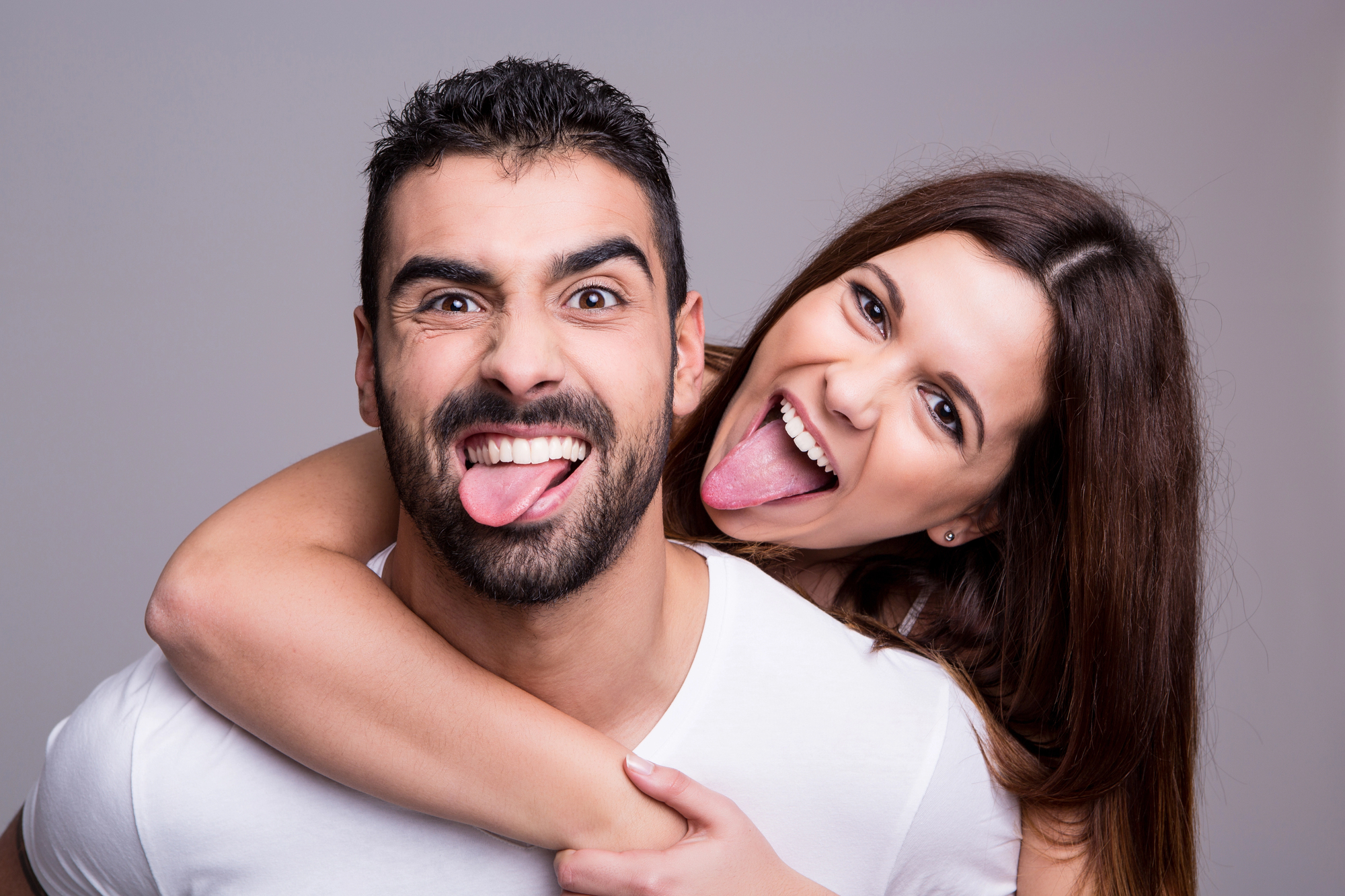 A man and a woman are posing playfully. The woman is piggybacking on the man, and both are sticking out their tongues, smiling widely. They are wearing casual white T-shirts and have dark hair, with the background being a plain, light color.