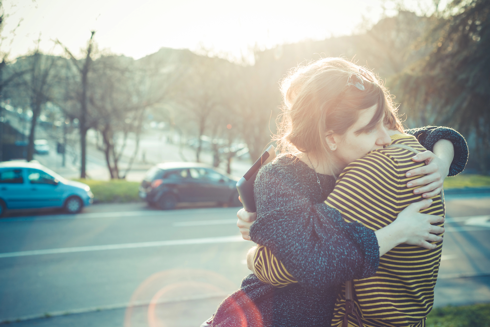 Two people hug warmly on a sunlit street. One person has sunglasses resting on her head and wears a dark sweater, while the other wears a striped shirt. Cars and trees are visible in the blurred background, giving a sense of a peaceful, urban setting.