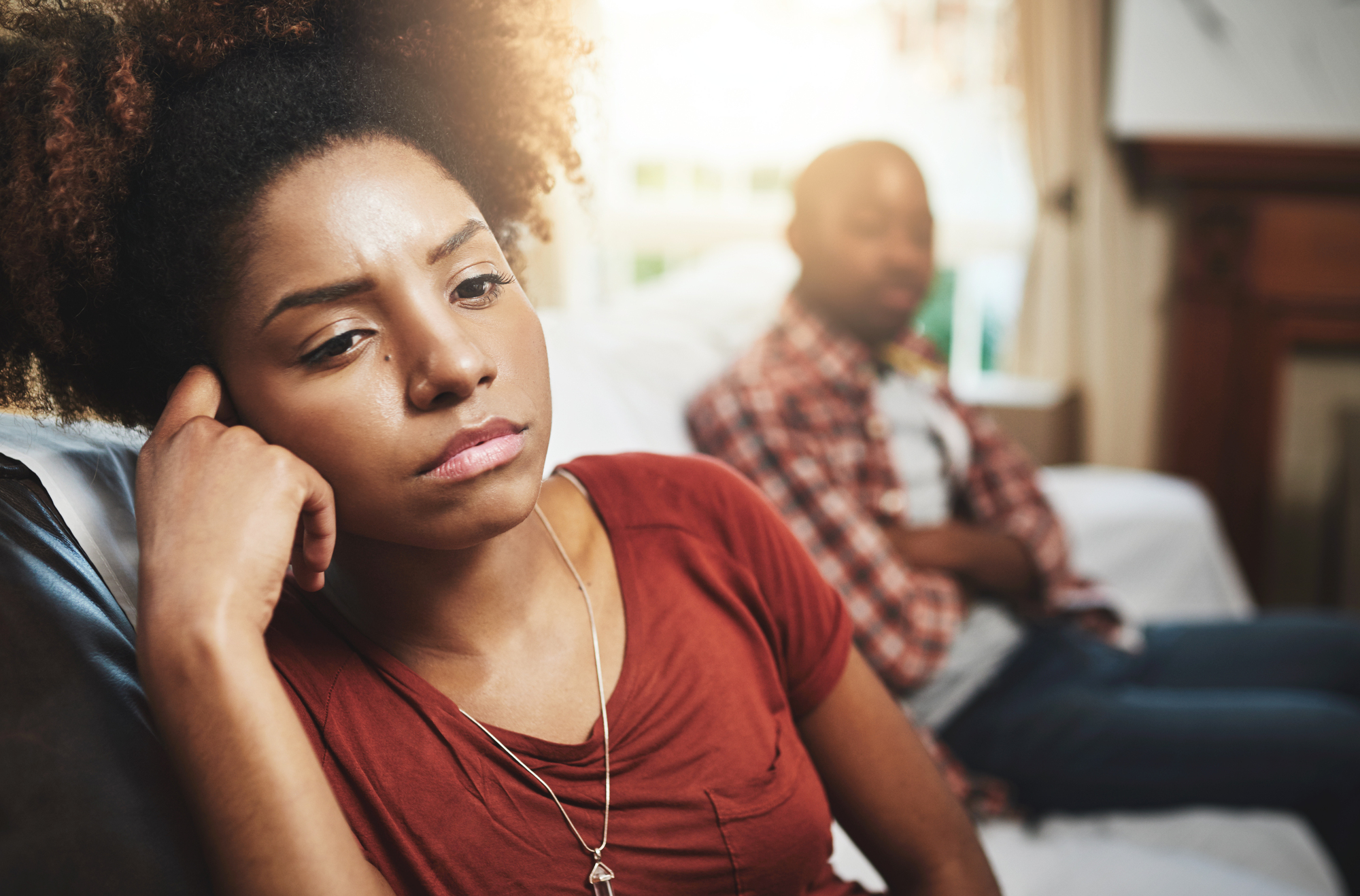 A woman with curly hair and a pensive expression sits on a couch, propping her head with one hand. She wears a red shirt and a necklace. In the background, a man in a plaid shirt and jeans also sits on the couch, looking away. The mood appears tense.