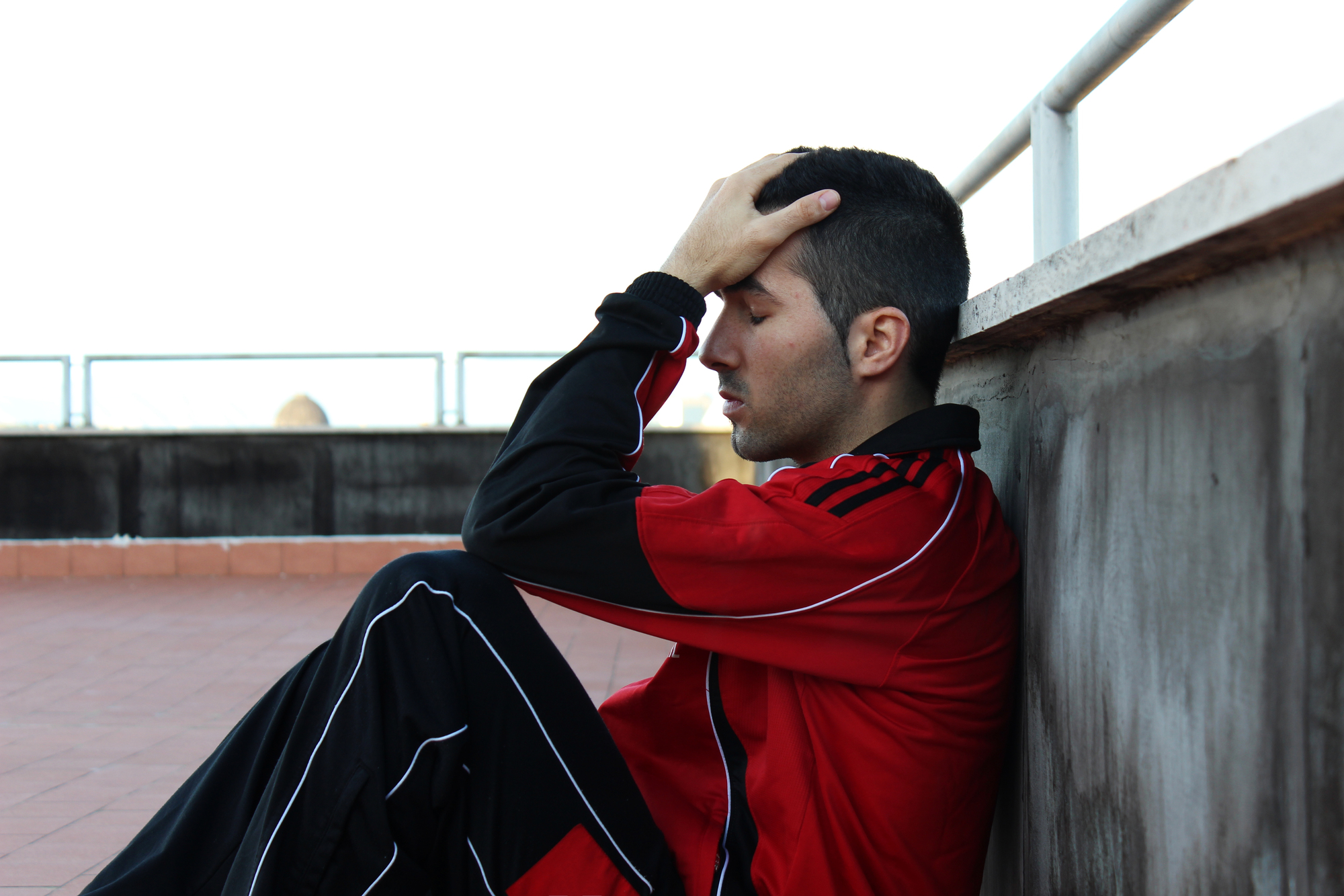 A person wearing a red and black tracksuit is sitting on the ground, leaning against a wall with legs bent and eyes closed. One hand is resting on their forehead, suggesting they are deep in thought or experiencing stress. The background is a rooftop with railings.