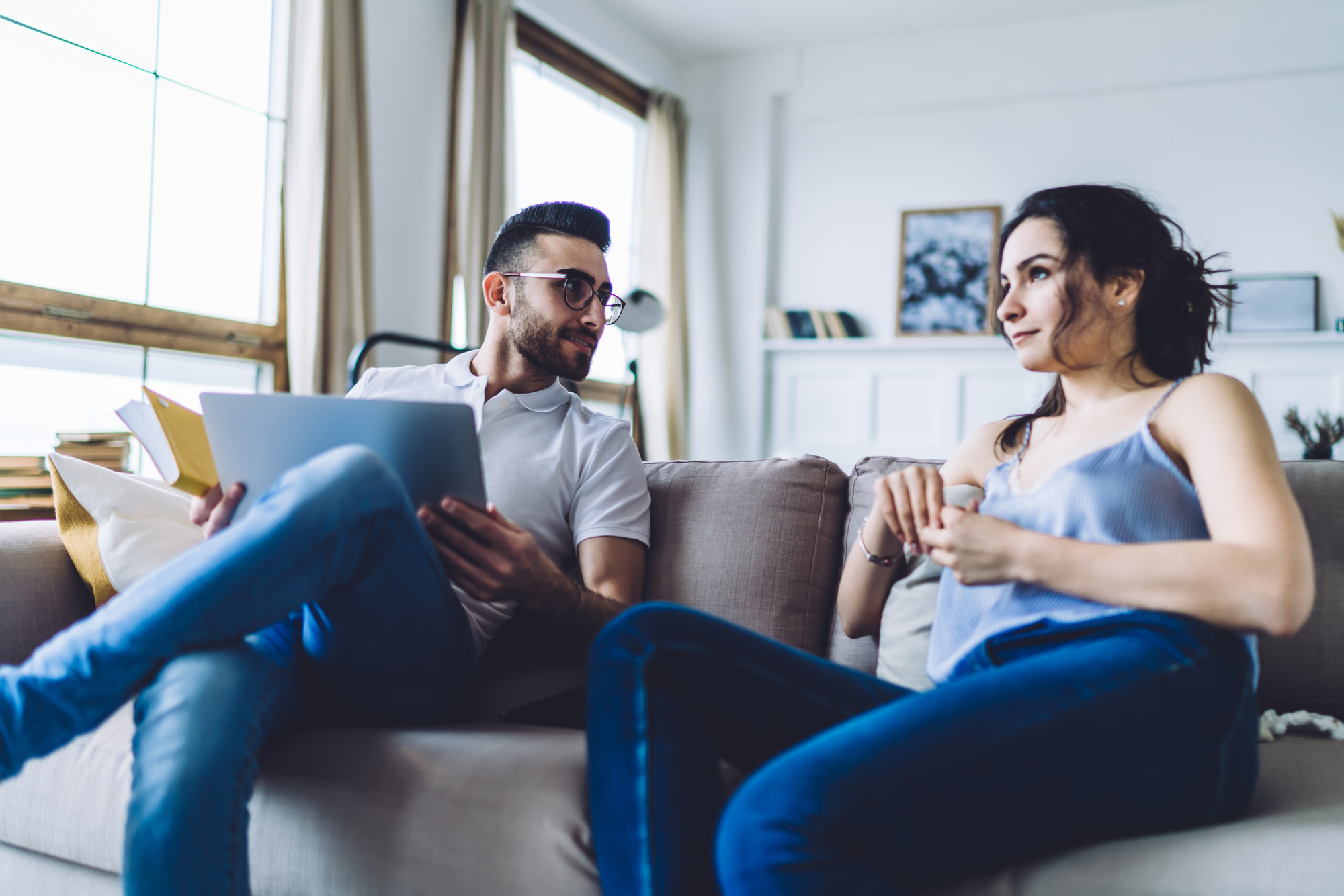 A man and woman are sitting on a couch in a bright living room. The man, holding a tablet and paperwork, looks at the woman attentively. The woman, in a blue tank top and jeans, is facing him with a relaxed demeanor. Sunlight streams through a large window.