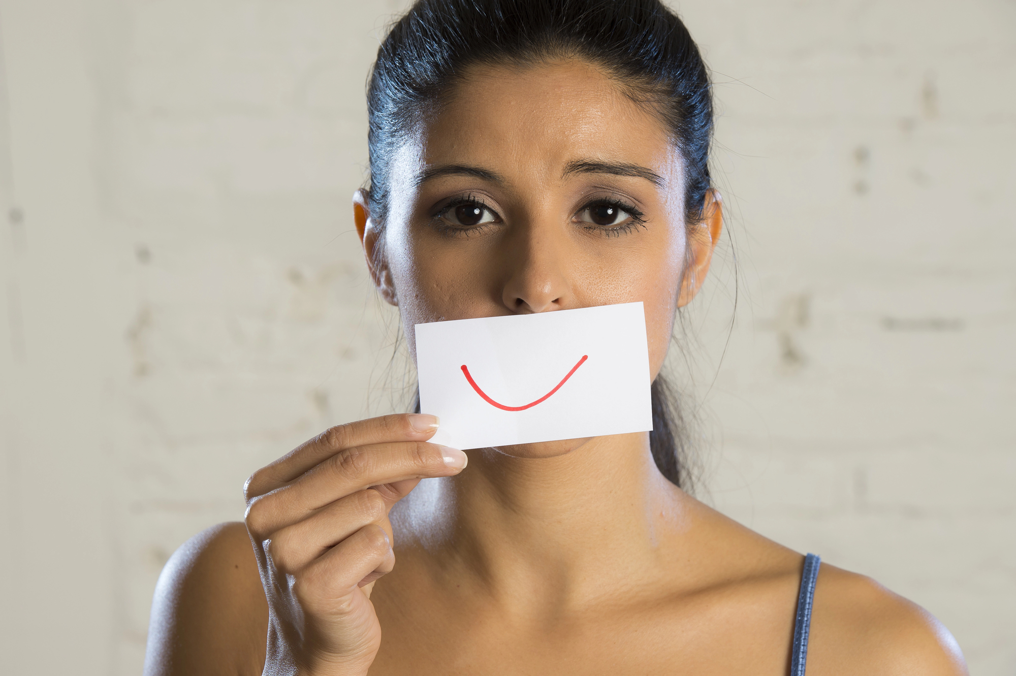 A woman with dark hair pulled back is holding a small white card in front of her mouth. The card has a red smile drawn on it, concealing her actual mouth. The background is blurred and appears to be a light-colored, textured wall.